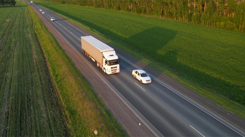 Ein weißer Lkw fährt über eine Landstraße, rechts und links von der Straße sieht man grüne Felder. Es sind weitere Fahrzeuge vor und hinter dem Lkw zu sehen. 